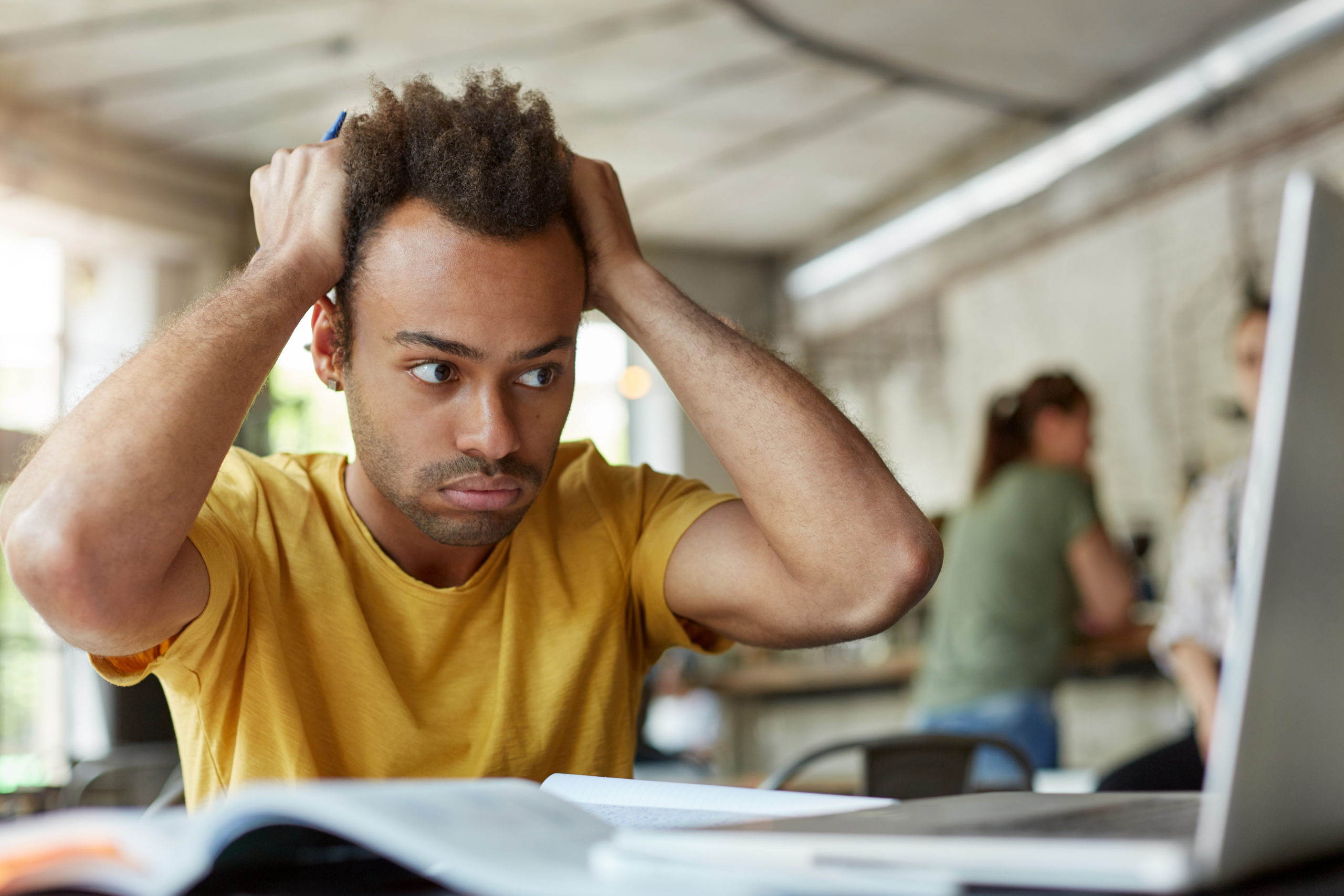 Stressed young African American student of school of economics feeling frustrated while working on diploma project, sitting at coworking space in front of open laptop computer, holding head with hands